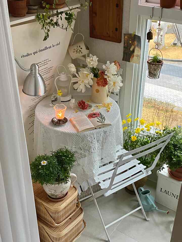 ホワイトとナチュラルカラーのベランダ、レースのテーブルクロスとヴィンテージチェア、キャンドルや多様な植物が配置された雰囲気のある空間