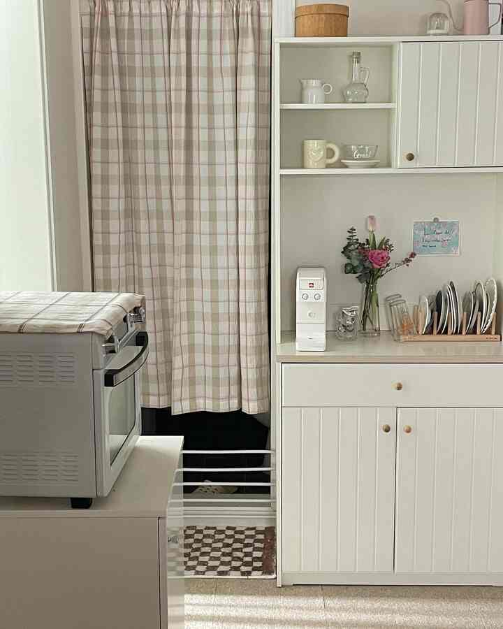 Cozy kitchen space with beige checkered curtains and white storage cabinets, featuring a coffee machine and plate rack, creating a home cafe vibe