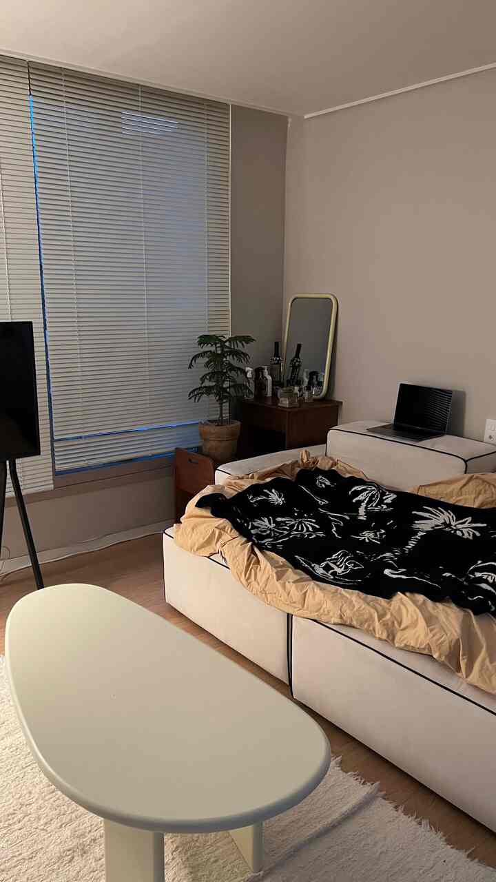 A cozy living room with white and brown tones featuring a cream sofa, oval coffee table, and aluminum blinds on the window
