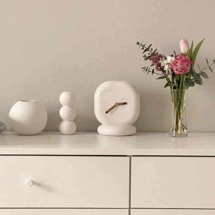 A calm beige wall with white dresser showcasing white ceramic objets and a vase with flowers in a cozy bedroom setting
