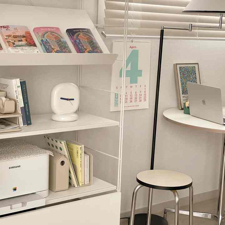 White-toned home office space featuring a round desk, stool, clean shelves, and a small clock with simple decor