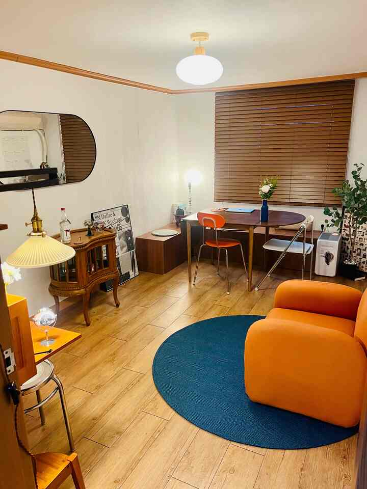 Natural wood tone studio living room featuring vintage furniture, an orange armchair, and a round blue rug creating a warm atmosphere