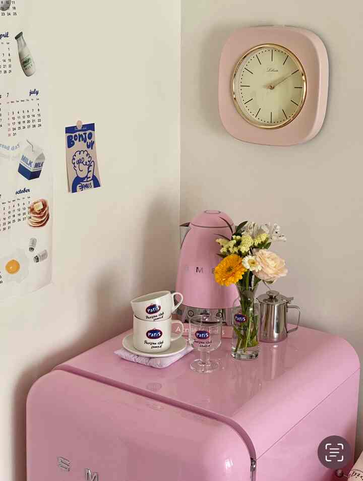 ピンクと白のトーンのキッチンコーナー、ヴィンテージスタイルのピンクのSMEG冷蔵庫と電気ケトル、花瓶に生けられた花がある雰囲気のあるインテリア