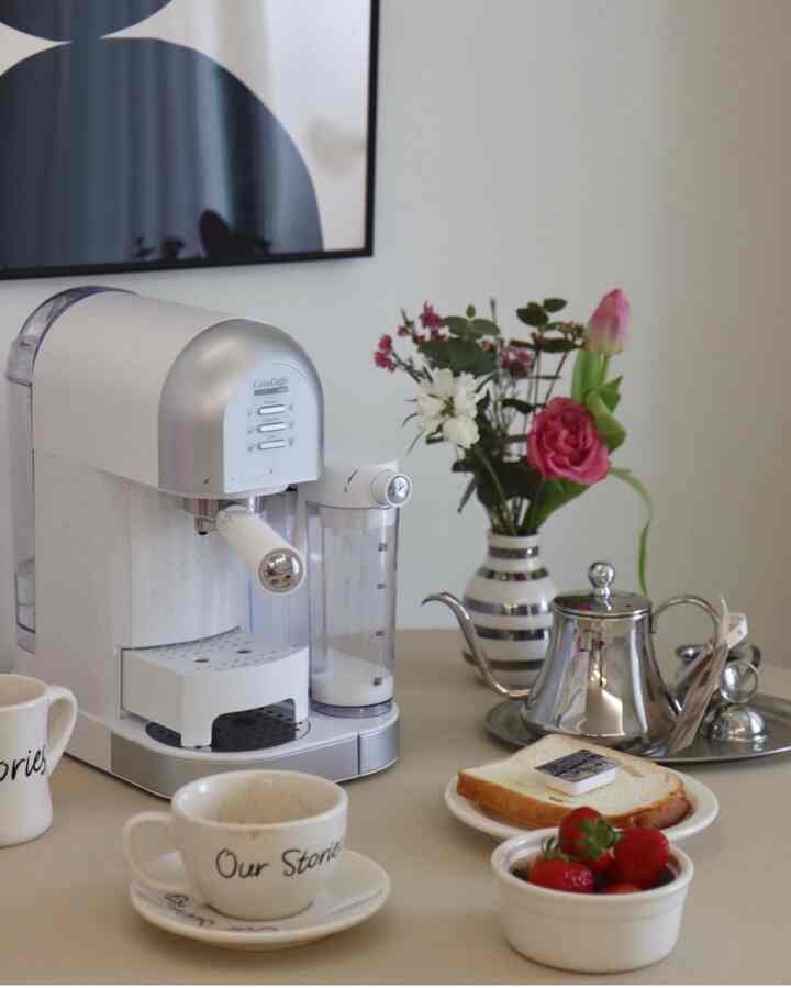 White and silver toned home cafe space featuring a coffee machine, mugs, vase with flowers, and dessert plate creating a cozy atmosphere
