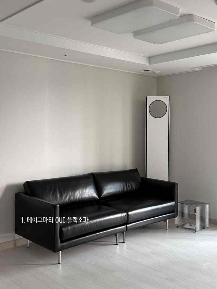 グレー調の壁と床が広がるリビングに、中央にブラックレザーのソファ、メタルワイヤーのサイドテーブル、右側に縦型エアコンが配置されたミニマルなインテリア