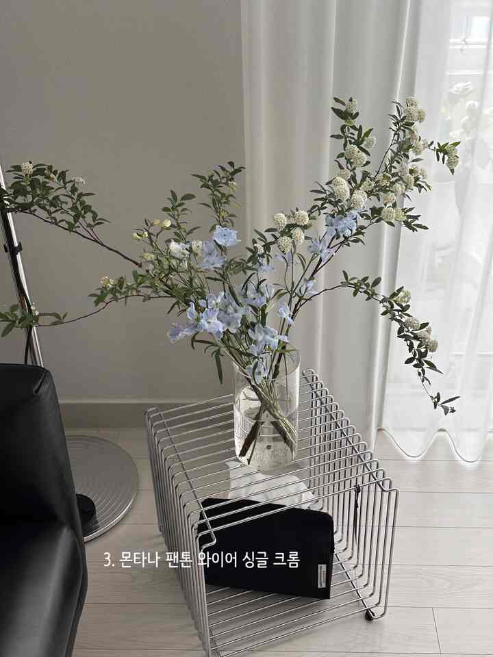 Modern and minimal living room with white walls and floor, chrome wire nightstand, transparent glass vase with flowers, and black sofa armrest