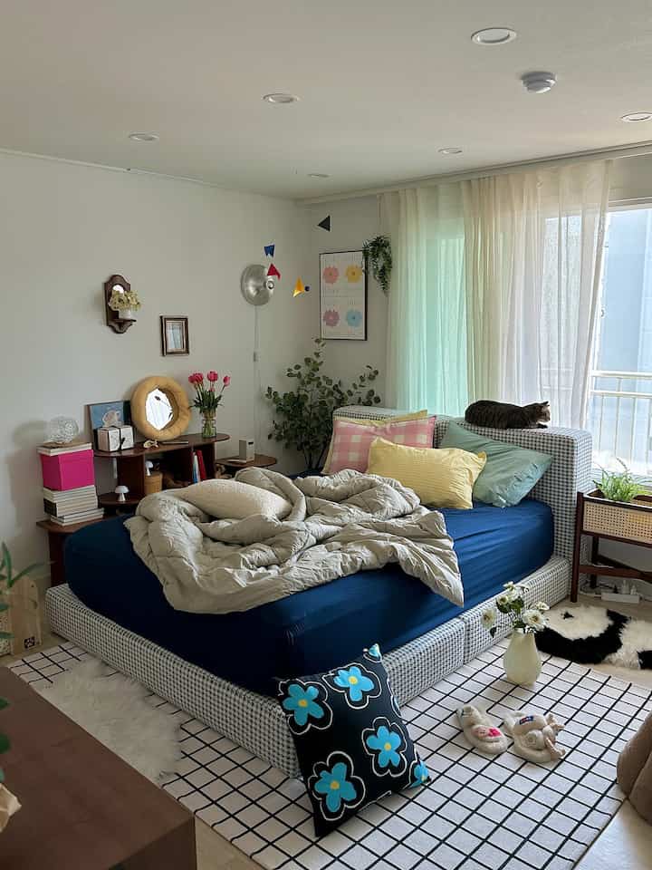 柔らかなナチュラルカラーの寝室、ファブリックベッドにカラフルなクッション、猫がくつろぐ印象的な空間
