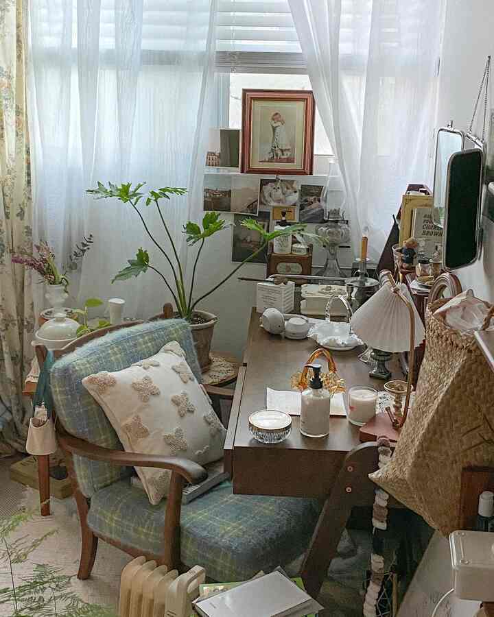 A natural-toned small room featuring curtains by the window, plants, a desk, and an armchair creating a cozy home office space