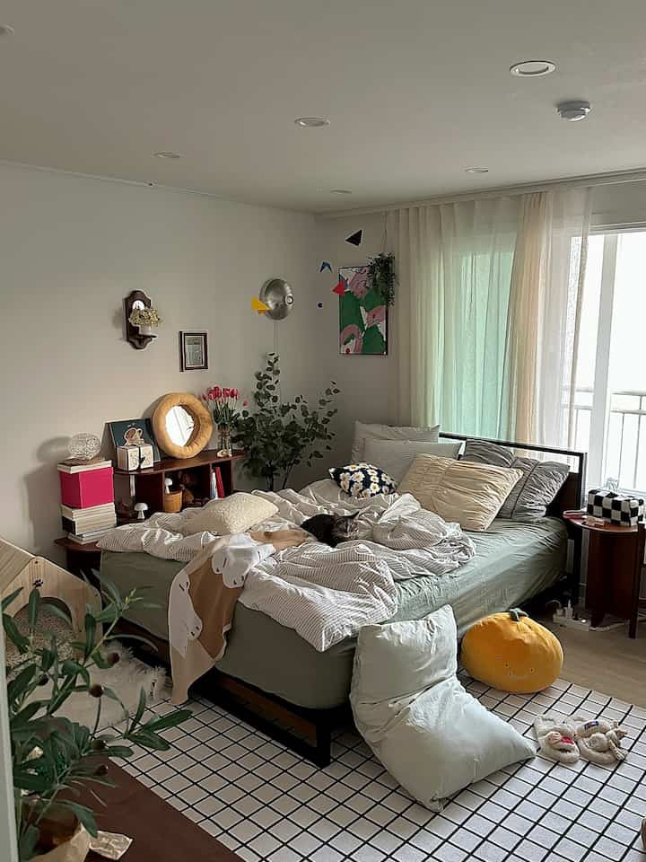 白い壁とカーテンのある寝室、花や植物が置かれた棚と居心地の良いベッドが調和する印象的な空間