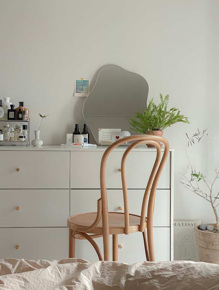 白と木目調の寝室、チェストと個性的な鏡、植物が配置されたシンプルで自然な雰囲気のインテリア