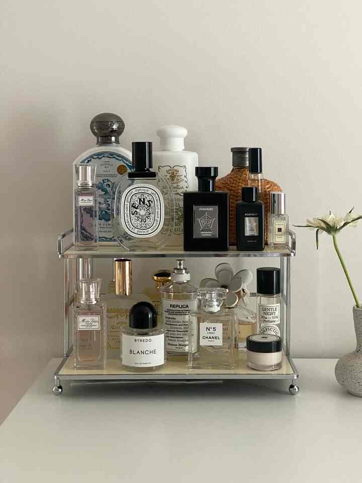 A white wall and table surface hold a two-tier metal tray featuring various neatly arranged perfume bottles, creating a tidy vanity accessory space.