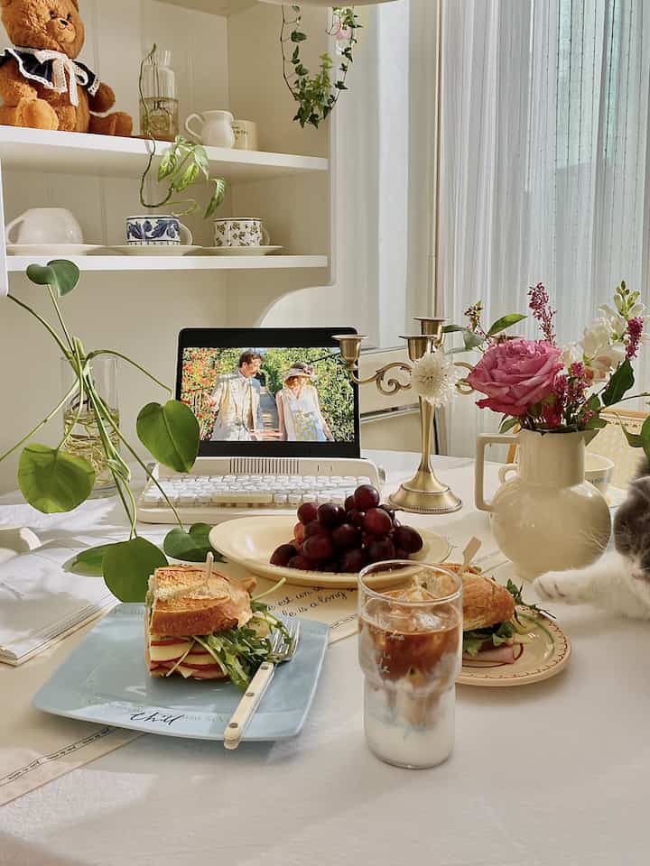 White and beige toned dining table space featuring sandwiches, coffee, a flower vase, and a cat's paw creating a cozy home cafe atmosphere