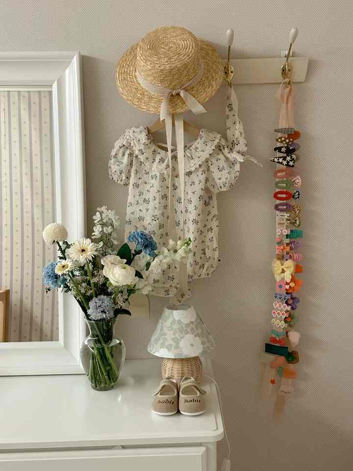 Natural-toned kids' room with white walls and furniture featuring child’s dress, hat, vase with flowers, lamp, and neatly organized hairpins