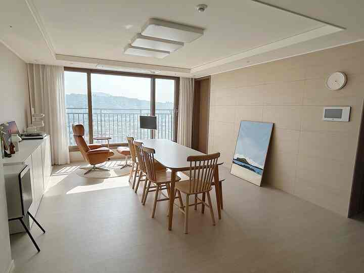 Bright natural-tone apartment living room featuring a Standby Me smart screen TV, single recliner, and wooden dining chairs creating a spacious calm atmosphere