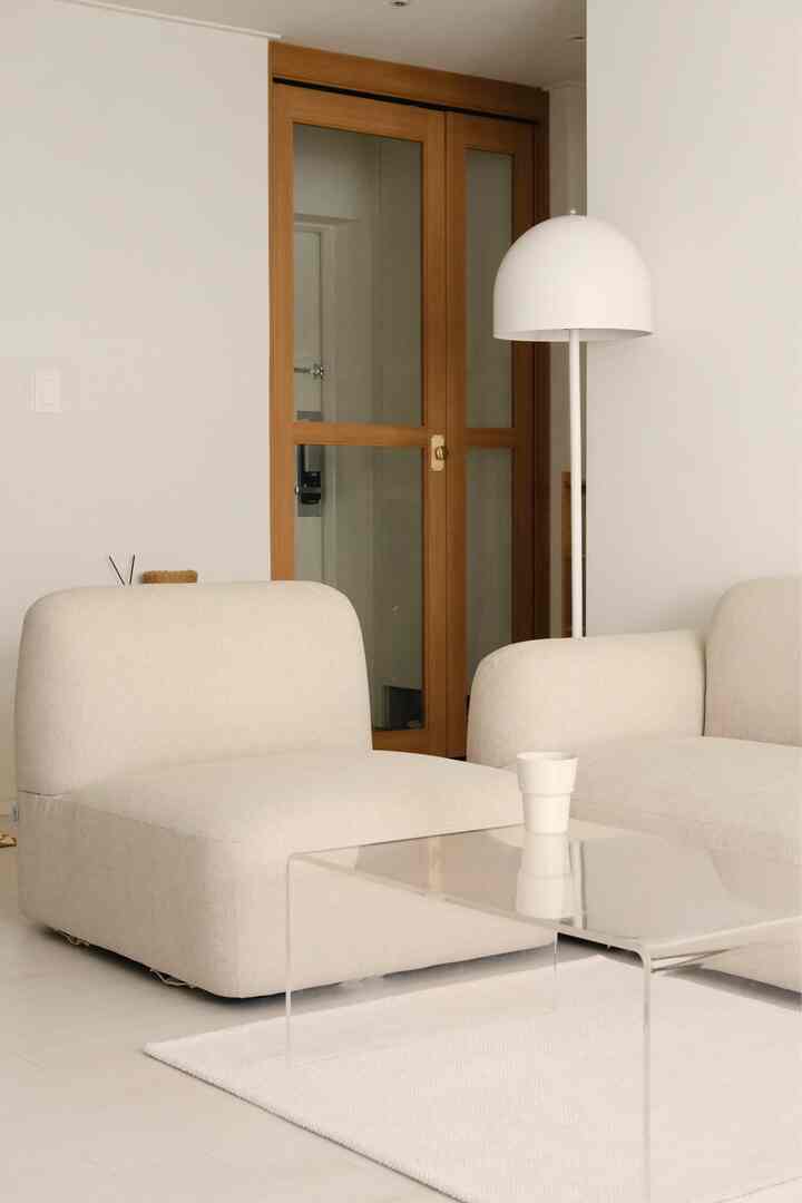 A white and beige toned living room featuring modular sofas and a transparent acrylic coffee table with a simple, natural atmosphere