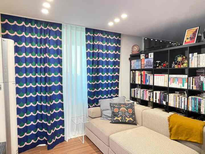 Living room with blue and beige tones, featuring a bookshelf as space divider, comfortable sofa, and vivid patterned curtains creating cozy atmosphere
