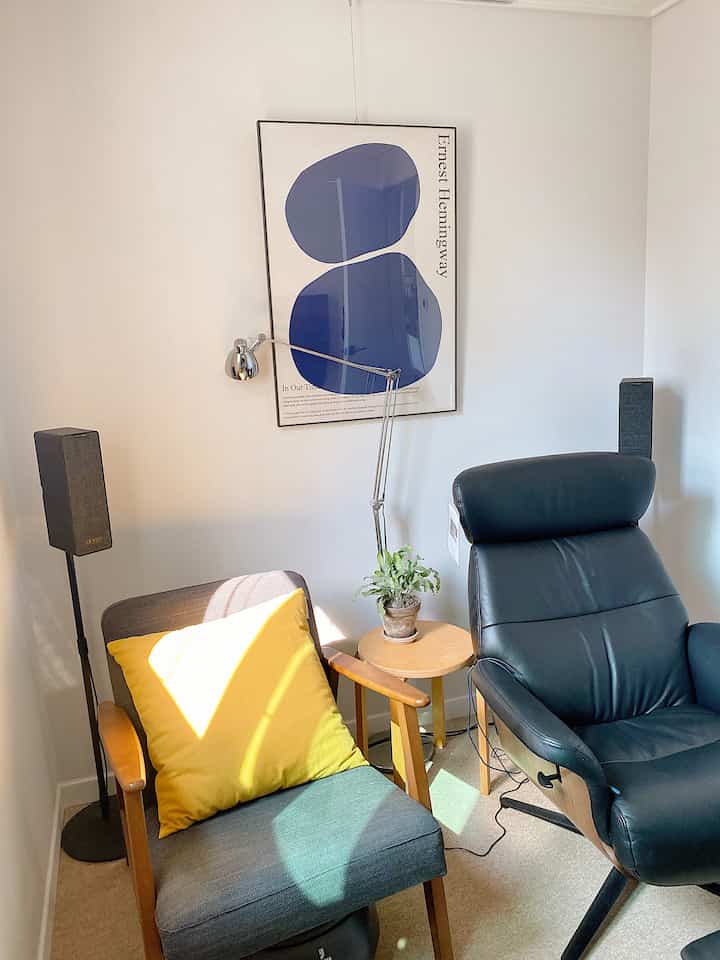 Bright white-walled small media room with brown wood round table, black recliner, and gray armchair with yellow cushion