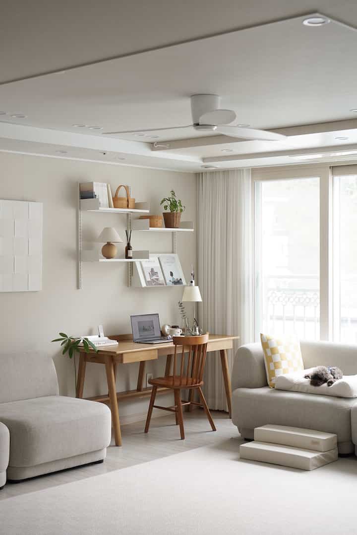 Beige and wood-tone living room and home office featuring a wooden desk, sofas, and a dog resting, creating a cozy atmosphere