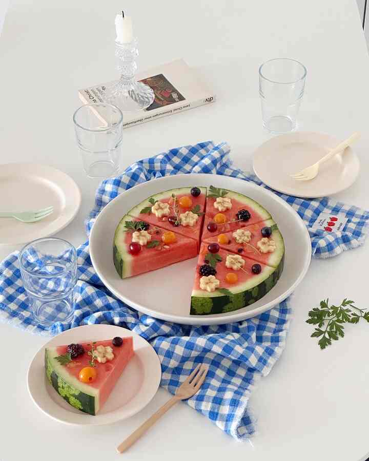 Minimal home party dining table with white surface, blue checkered cloth, and fruit-decorated watermelon pizza centerpiece