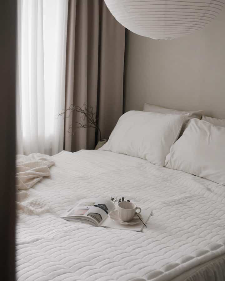 ホワイトとベージュの色調の寝室、白い寝具と枕、ベージュのカーテンが配置された自然で落ち着いた雰囲気のあるインテリア