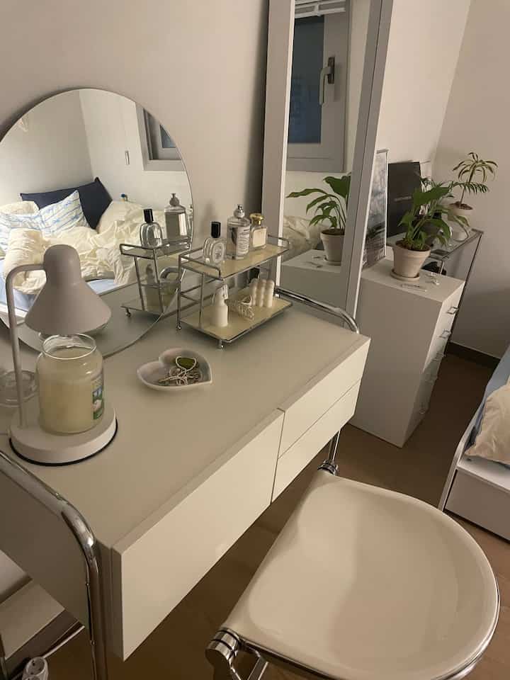 A simple bedroom with white and natural tones featuring a vanity with mirrors and plants, creating a cozy atmosphere