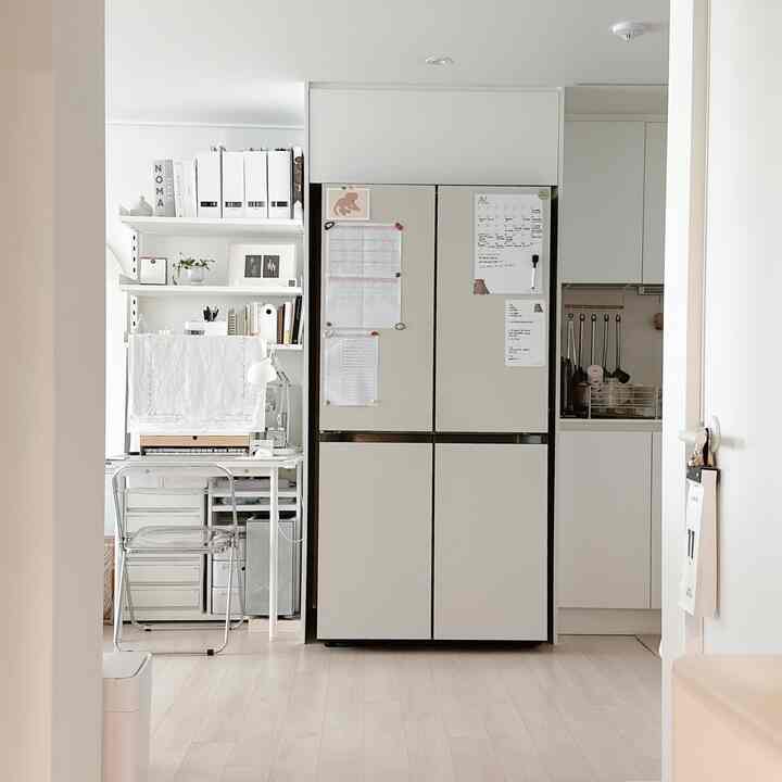 Modern kitchen and home office space featuring beige refrigerator, white wall shelves, and transparent chair
