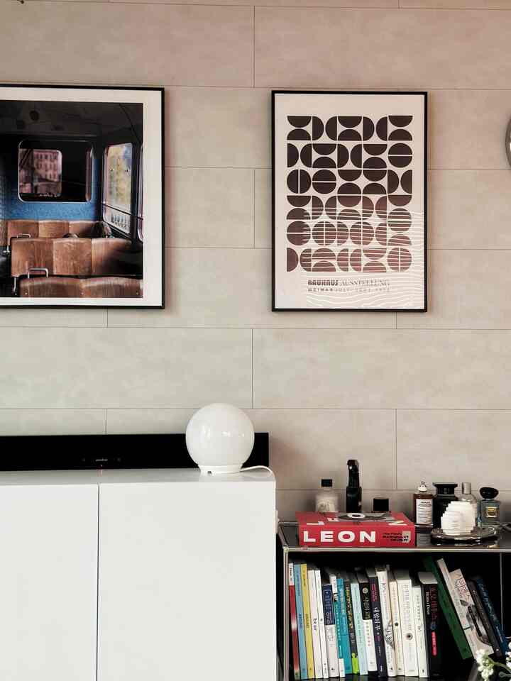 A simple living room with white and beige tones featuring two picture frames on the wall and a bookshelf and white cabinet at bottom right