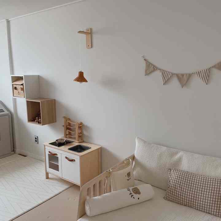 ナチュラルカラーのワンルームで、赤ちゃんと暮らすためのソファベッドと小さなキッチントイが配置された温かみのあるインテリア