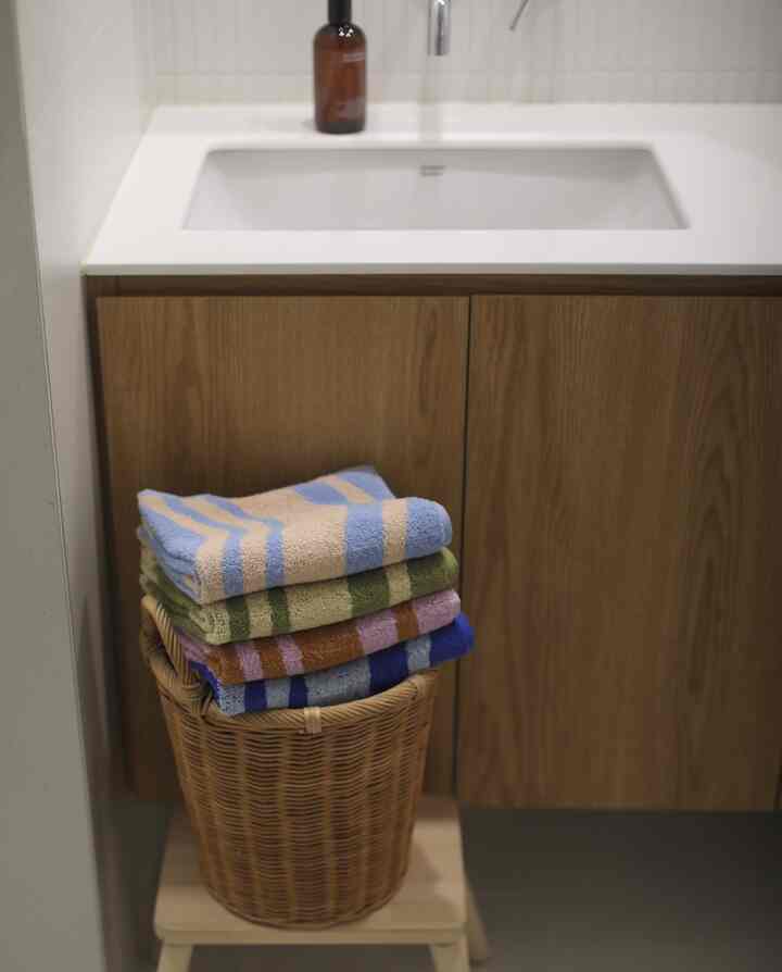 ナチュラルな木目調の収納棚と白い洗面台がある清潔感のある浴室、カラフルなストライプ柄のタオルが籐のバスケットに整然と積まれている
