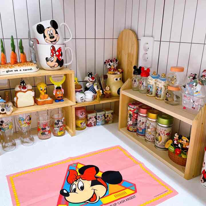 A bright kitchen space dominated by pink and wood tones, featuring neatly arranged Disney character mugs and wooden storage shelves creating a charming atmosphere