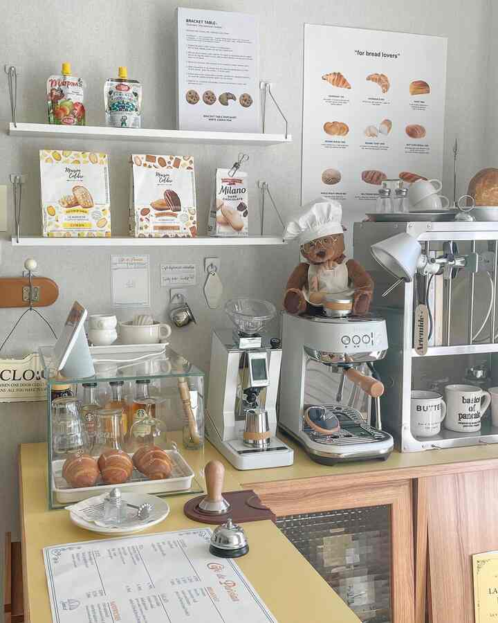 A white-walled kitchen space featuring a yellow counter with coffee machines and bakery items, creating a cozy home cafe atmosphere
