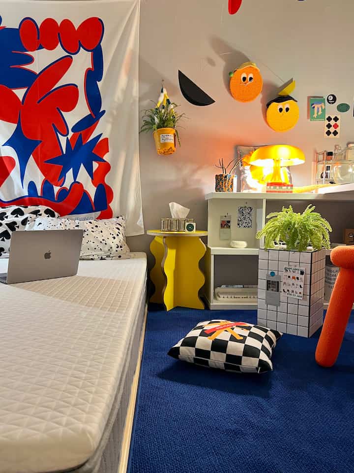 黄色のアクセントと青いラグが映える寝室空間、白いマットレスと黄色のサイドテーブル、テーブルランプが配置された雰囲気のあるインテリア