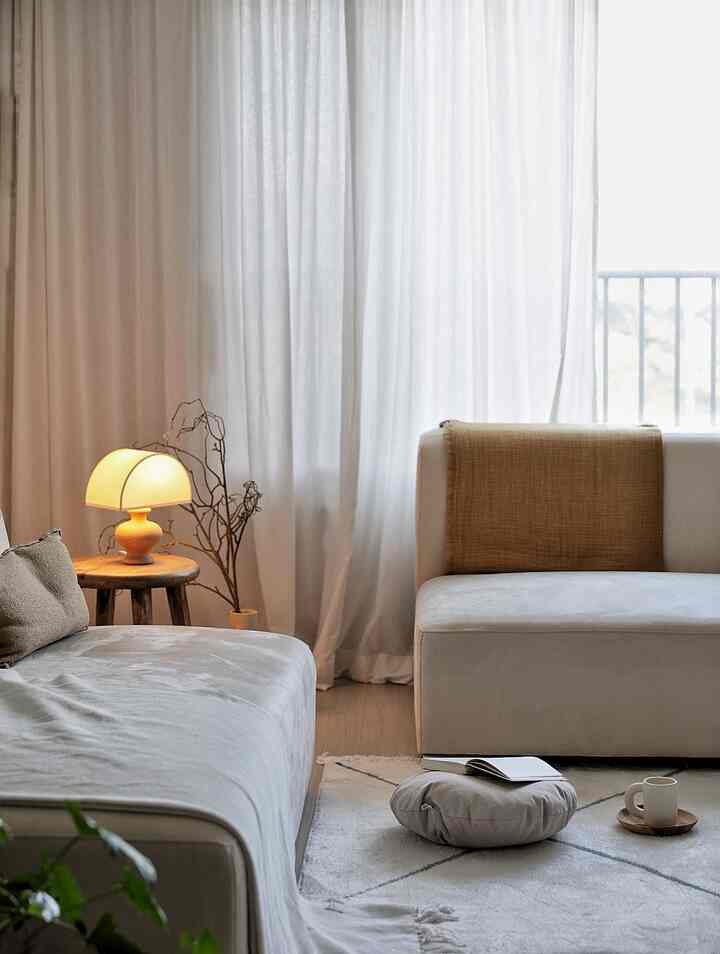 A natural tone living room featuring beige sofas, white curtains, wooden stool, and warm table lamp creating a cozy atmosphere