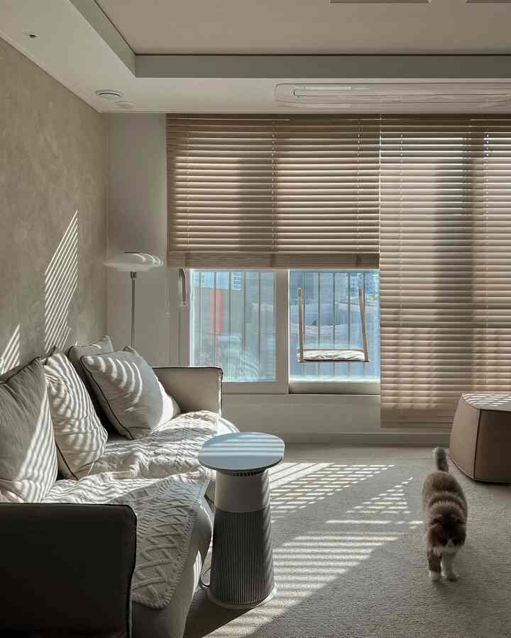 Beige and white toned living room featuring large windows with blinds, sofa, air purifier, and a cat in cozy atmosphere