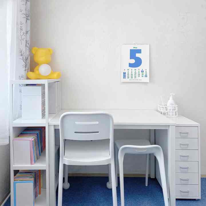 A white and blue toned kids' room featuring a large white desk with two chairs centrally placed and an organized bookshelf on the left, giving a clean atmosphere