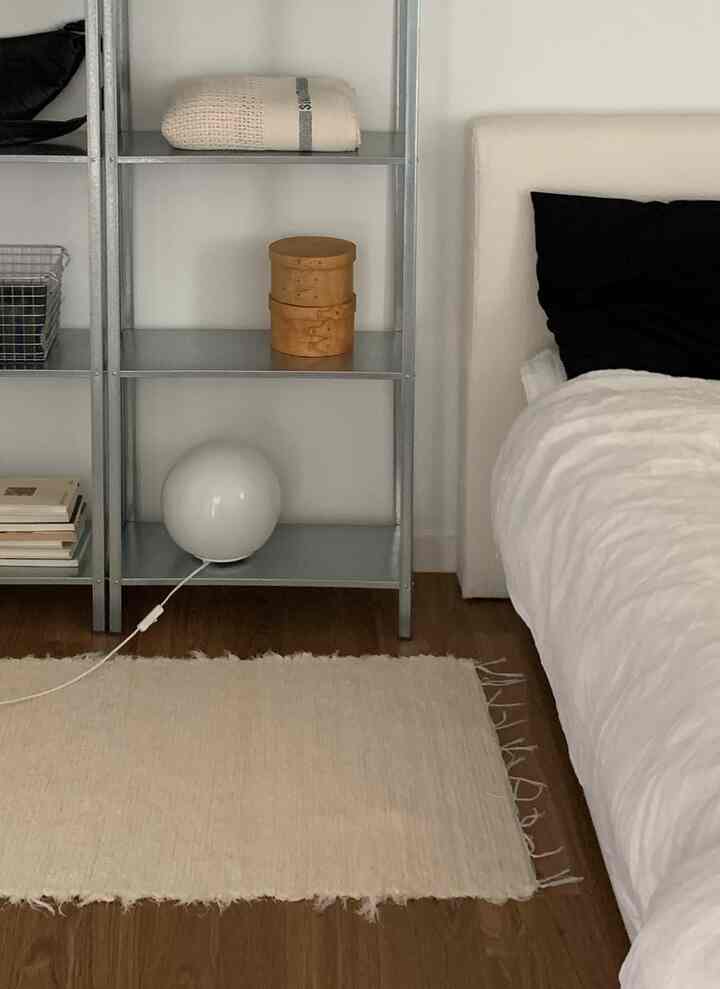 White and brown toned bedroom featuring a bed with white linens, beige rug on wooden floor, metal shelving unit and a spherical floor lamp creating a calm atmosphere