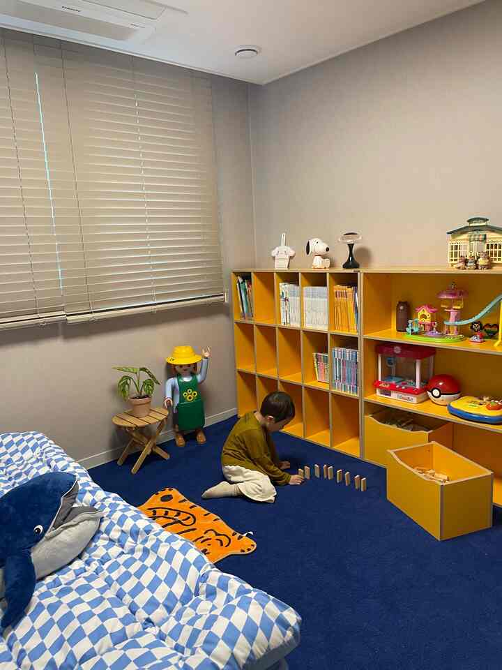 ネイビーブルーのラグと黄色の子供用本棚が配置された子供部屋、中心右寄りで遊ぶ子供の様子