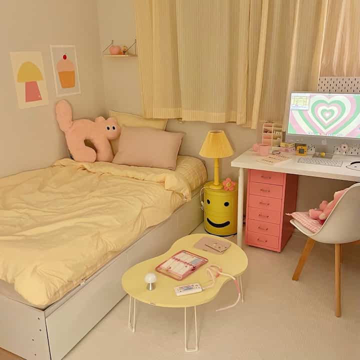 パステル調のワンルーム寝室、黄色い寝具とピンクの引き出し付きデスクが配置された雰囲気のある空間