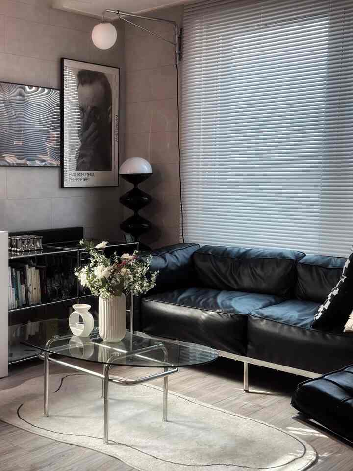 Modern natural-toned living room featuring black leather sofa, glass coffee table, and aluminum blinds creating a cozy atmosphere