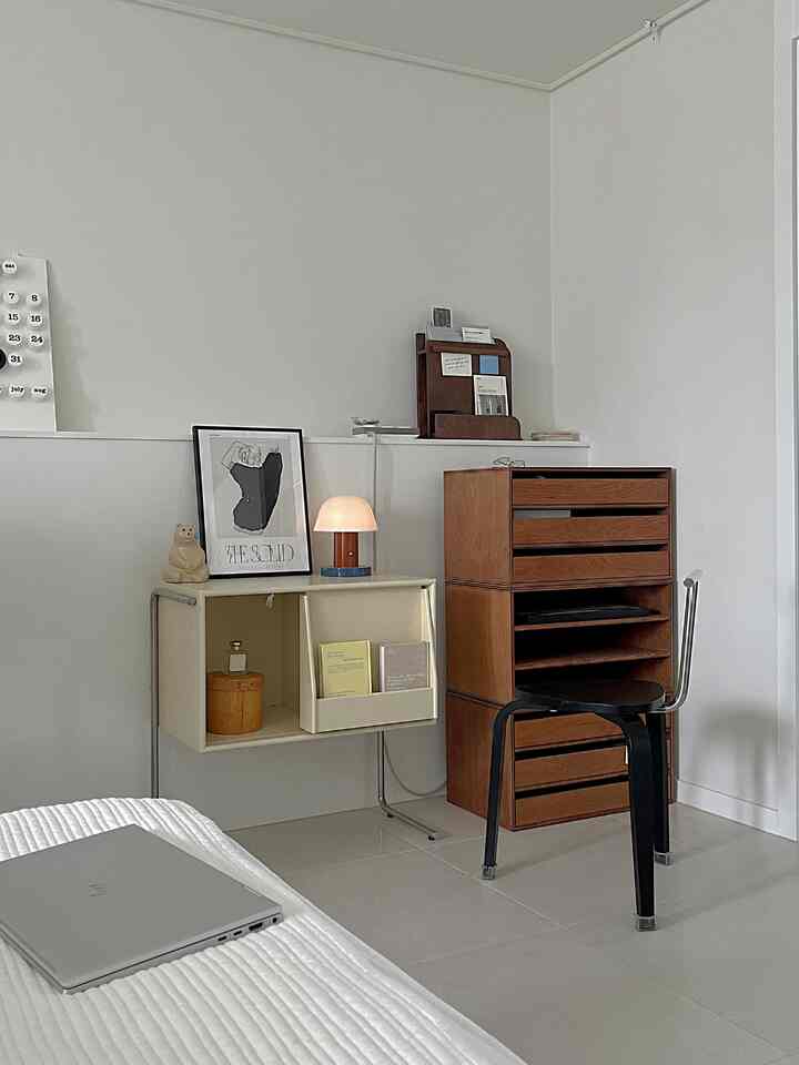 白い壁と床、茶色の木製家具とスツールが配置されたミニマルなワンルームのホームオフィス兼寝室空間