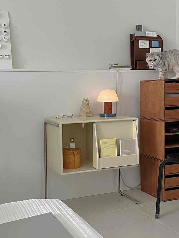 ナチュラルトーンの小さな部屋、壁にアイボリーの棚とテーブルランプがあり、猫がいる印象的な空間