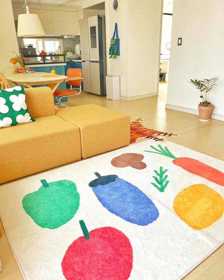 Natural-toned living room featuring bright yellow sofa and orange dining chairs, accented by a playful vegetable-patterned rug