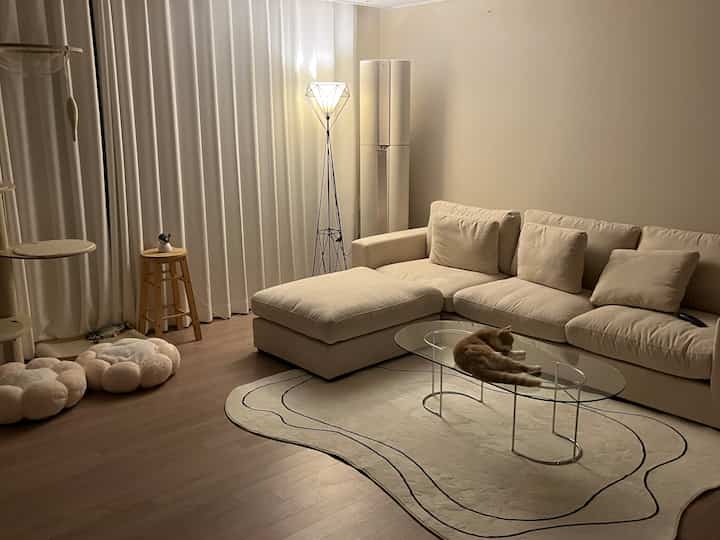 A simple living room in beige and wood tones featuring a sectional sofa, glass coffee table, and a cat lounging