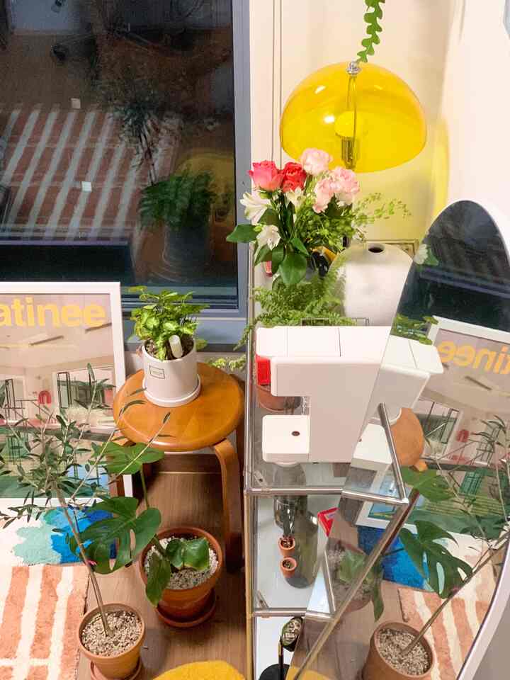 黄色のトーンが特徴のワンルーム、木製スツールと透明なモジュール棚、全身鏡、多数の植物と花瓶が配置された雰囲気のあるモダンなミッドセンチュリー・モダンインテリア