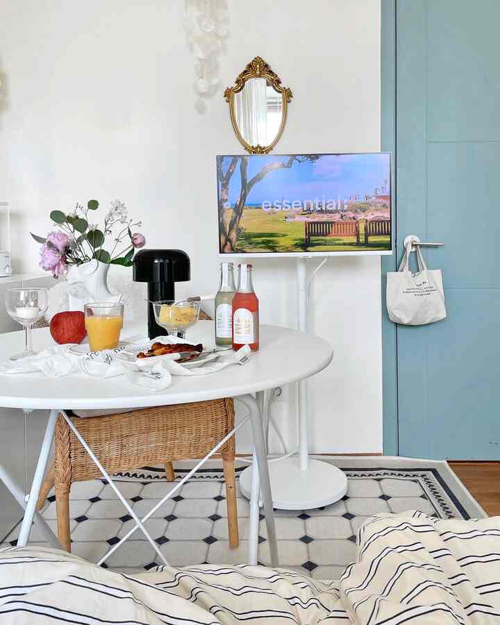 白と青のトーンが特徴のダイニングルーム、中央に丸テーブルとラタン製ダイニングチェアが配置され、壁には金色の鏡とテレビが設置されたシンプルな空間