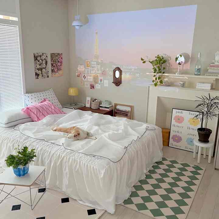 白とピンクのアクセントがある寝室、中央に猫が横たわるベッドと右側にダイヤモンド柄のラグが配置された雰囲気のあるインテリア