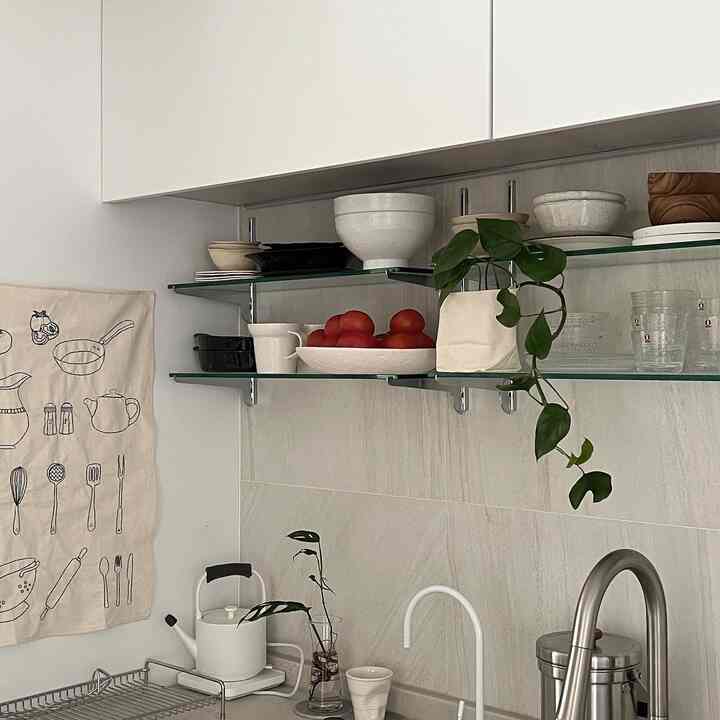 White-toned kitchen space featuring glass shelves with kitchenware and plants, creating a clean and natural atmosphere