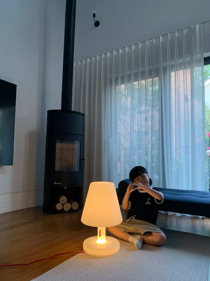 白い壁とナチュラルな木製の床のリビング、暖かい光のフロアランプとネイビーのソファ、子供が座っている空間