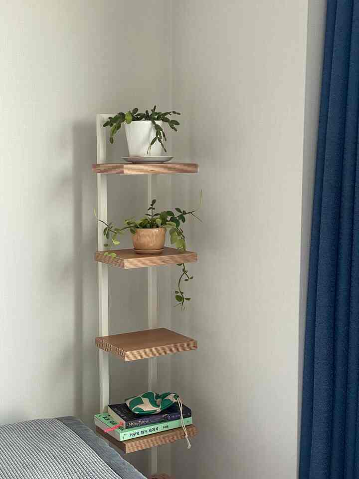 白い壁とネイビーカーテンの寝室コーナー、植物と本が置かれた木製のコーナー棚があるナチュラルモダンな空間