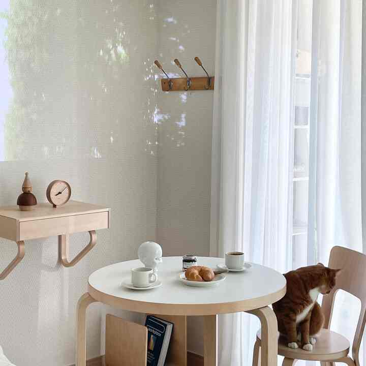 A small white and wood-toned dining space featuring a round table, chair with a cat, and sheer curtains, presenting a minimal and natural ambiance